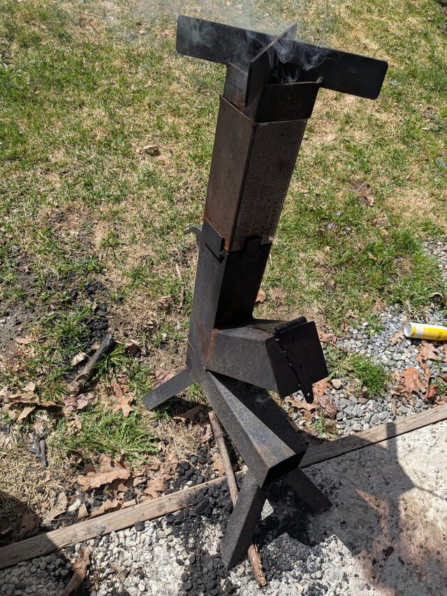 Welded steel rocket stove prototype — vertical burn chamber with a fuel-feed shelf, tested outside the shop